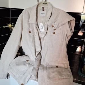 Beige jacka från Cosy Mode - Säljer en stilren beige jacka från Cosy Mode. Jackan har långa ärmar och knappar framtill. Den är tillverkad i linne och har en snygg innerfoder med mönster. Perfekt för vår och sommar!