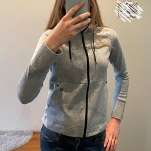 Grå hoodie från SOC - En fin grå hoodie från märket SOC i storlek XS. Passar perfekt nu på våren både som ytterplagg men också under andra jackor. Yttertyget är i bomull men den är vadderad med polyester.(Ganska tight passform för mig, men jag har egentligen storlek S) Har lite noppriga partier kring ärmar och även vid fållen men det är knappt märkbart. 
