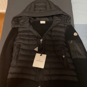 Svart dunjacka från Moncler - Snygg svart dunjacka från Moncler med stickade ärmar och quiltad kropp. Jackan har en dragkedja framtill och är en snygg jacka för hösten/sommarn