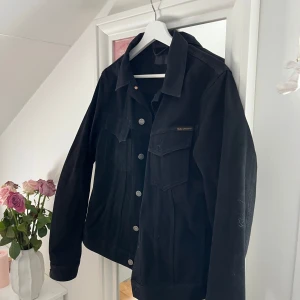 Svart jeansjacka från Nudie Jeans - Säljer en stilren svart jeansjacka från Nudie Jeans. Jackan har knappar framtill och två bröstfickor med lock. Perfekt för en casual look och passar till det mesta. 🖤