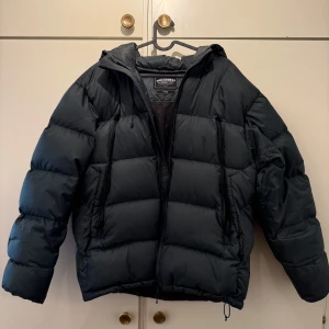 Grön pufferjacka från Frizmworks - Säljer en hrön pufferjacka från Frizmworks i storlek L. Jackan har en dragkedja framtill och är långärmad med en mysig huva. Perfekt för kyliga dagar.