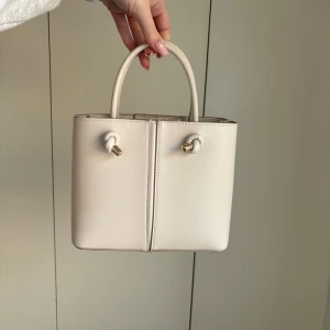Handväska från Zara - En ljusbeige handväska från Zara. Superfin men ej använd. Finns ett långt band till som man kan ta av om man vill. Fack i mitten med dragkedja och stängning med knapp upptill.  Mått: 23x19x10