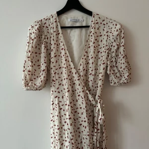 Vit omlottklänning med körsbärsmönster från ADOORE - Säljer en söt vit omlottklänning från ADOORE med röda körsbärsmönster. Klänningen har korta puffärmar och ett knytband i midjan för en smickrande passform. Perfekt för somriga dagar!