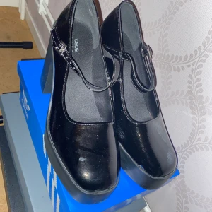 Svarta Mary Jane skor med klack från ASOS - Snygga svarta Mary Jane skor från ASOS med hög klack och justerbar rem över foten. Skorna har en glansig finish och en rundad tå, perfekt för en stilren look. Endast använda 2 gånger. Är dessutom i nyskick i storlek 39, passar 40 också! Köpta för 380kr! Pris kan diskuteras