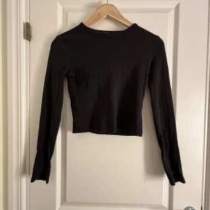 Svart ribbad topp från Bershka - Säljer en svart ribbad topp från Bershka. Den har en croppad design med långa ärmar, perfekt för en stilren look. Passar bra till både jeans och kjol. Perfekt för höst och vår!