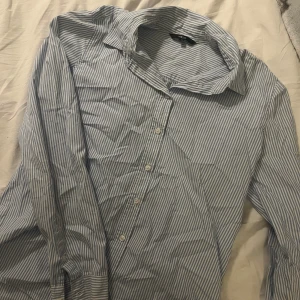 Blå- och vitrandig skjorta från Ellos - Säljer en klassisk blå- och vitrandig skjorta från Ellos. Skjortan har långa ärmar och knappar framtill. Perfekt för en stilren look.