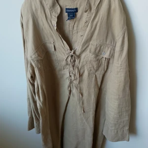 Ny klänning från GANT - Säljer en stilren beige klänning från GANT med långärmad design och knytning framtill. Klänningen har två bröstfickor med knappar och en avslappnad passform. Perfekt för en avslappnad och chic look.