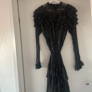 Svart långklänning Dodo BarOr  - Elegant svart långklänning med volanger och guldiga ränder. Klänningen har långa ärmar och ett knytband i midjan för en smickrande passform. Perfekt för en festlig kväll.