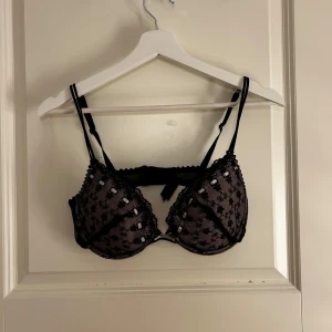 Svart spets-bh med rosett - Elegant svart bh med spetsdetaljer och en söt rosett framtill. Den har justerbara axelband och en dekorativ kant med små prickar. Perfekt för en stilfull look.