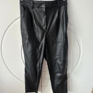 Svarta skinnbyxor från H&M - Snygga svarta skinnbyxor från H&M i storlek 42. Byxorna har en klassisk design med hög midja och raka ben. Perfekta för en stilren look. Tillverkade i syntetmaterial som ger en läderliknande finish. Ett litet skav på baksida
