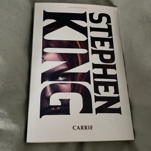 Carrie - Säljer boken 'Carrie' av Stephen King. En klassisk skräckroman om en tonårsflicka med telekinetiska krafter. Perfekt för dig som älskar spänning och skräck. Boken har en vit omslag med titeln i stora svarta bokstäver och en bild av en flickas ansikte.
