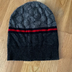 Grå och svart mössa med röd rand - Snygg och stilren mössa i grått och svart med en röd rand. Den har en ribbad kant som ger en skön passform. Perfekt för kyliga dagar.