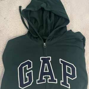 Grön hoodie från GAP - Säljer en snygg grön hoodie från GAP med dragkedja och stor logga på framsidan. Perfekt för en avslappnad stil. Den har en praktisk huva med dragsko.