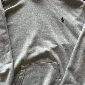 Grå hoodie från Polo Ralph Lauren - Säljer en stilren grå hoodie från Polo Ralph Lauren med det klassiska broderade logotypen på bröstet. Tröjan har en känguruficka och långa ärmar, perfekt för en avslappnad look. Passar utmärkt för kyligare dagar.