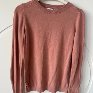 Rosa tunnstickad tröja med pärldetaljer - Säljer en söt rosa tunnstickad tröja med långa ärmar och fina pärldetaljer på ärmsluten. Perfekt för att ge en touch av elegans till din outfit. Tröjan har en rund halsringning och ribbade kanter.