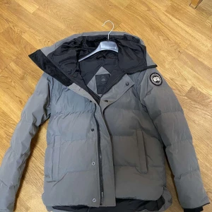 Grå macmillan f Canada Goose - Säljer en grå dunjacka från Canada Goose med deras ikoniska märke på ärmen. Jackan har en justerbar huva och dragkedja framtill. Perfekt för kalla dagar med sin varma och skyddande design. Storlek xs passar 165-170