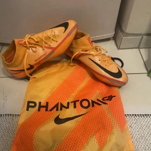 Orange fotbollsskor från Nike - Snygga och iögonfallande orange fotbollsskor från Nike med svarta och röda detaljer. Skorna har snörning och en modern design som passar perfekt för fotbollsplanen. Levereras med en matchande påse. Köpta för 2799kr och är som helt nya då jag använt dem 2 gånger.