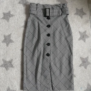 Rutig kjol med bälte från H&M - Snygg rutig kjol från H&M med knappar framtill och ett matchande bälte i midjan. Kjolen har en klassisk svartvit design och är perfekt för en stilren look. Den är knälång och har en elegant passform.