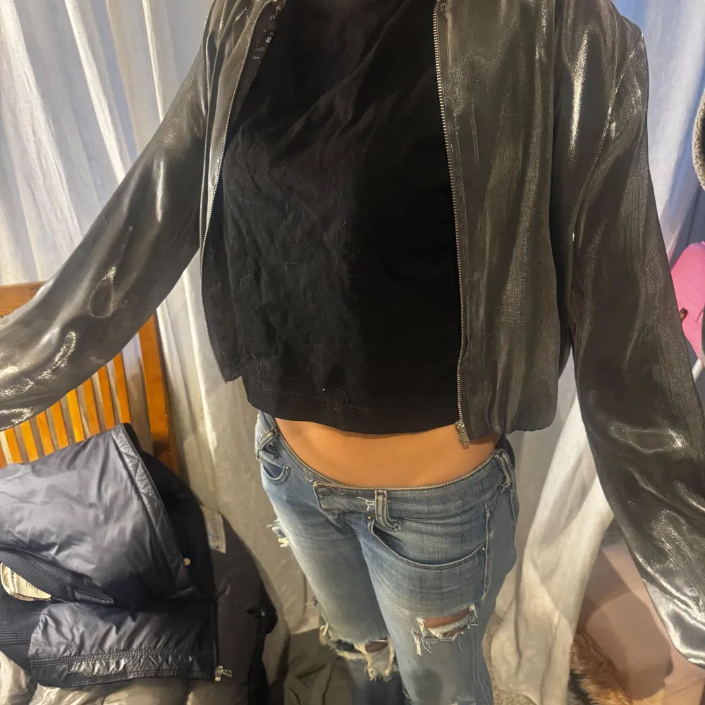 Snygg silverfärgad glittrig jacka med dragkedja framtill. Jackan har en croppad passform och långa ärmar, perfekt för en kväll ute. Den glänsande ytan ger en festlig touch.. Takit.