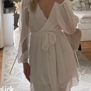 Vit klänning  - Säljer en söt vit omlottklänning med prickigt mönster. Klänningen har långa ärmar och ett knytband i midjan för en fin passform. Perfekt för sommarens alla tillfällen och studenten. 