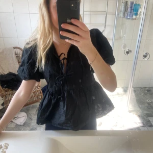 Svart blus med knytning - Säljer en svart blus med puffiga korta ärmar och knytning framtill. Blusen har en luftig passform och volangdetaljer vid midjan. Perfekt för en stilren look.