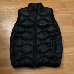  Peak Performance väst storlek 160 - Snygg svart dunväst från Peak Performance med dragkedja framtill. Perfekt för kyliga dagar och ger en sportig look. Västen har en quiltad design som ger extra värme och stil. Storlek 160. Priset kan diskuteras. 