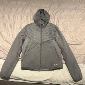 Grå Nike Windbreaker  - Säljer en grå vindjacka från Nike med huva och dragkedja. Jackan har långa ärmar och en stilren design med Nike-logga på framsidan. Perfekt för blåsiga dagar.