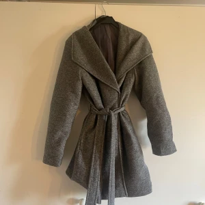 Grå kappa från Vero Moda - Snygg grå kappa från Vero Moda med knytbälte i midjan. Den har en elegant krage och stängs med tryckknappar. Perfekt för kyliga dagar eller vår morningar