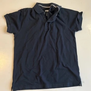 Mörkblå pikétröja från Helly Hansen - Säljer en stilren mörkblå pikétröja från Helly Hansen. Tröjan har korta ärmar och en klassisk krage med knappar. Perfekt för en avslappnad look.