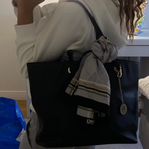 Svart axelväska från DKNY - Snygg svart axelväska från DKNY med en dekorativ scarf och guldfärgad kedjedetalj. Väskan har ett rymligt inre med DKNY-mönstrat foder och en praktisk dragkedja. 