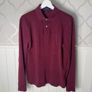 Långärmad pikétröja från Polo Ralph Lauren - Säljer en stilren vinröd pikétröja från Polo Ralph Lauren med klassisk krage och knappar framtill. Tröjan har långa ärmar och en broderad logotyp i blått på bröstet. Perfekt för en kyligare sommardag men ändå elegant look. Hör av er vid eventuella frågor! 