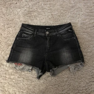 Svarta jeansshorts från Replay - Snygga svarta jeansshorts från Replay med fransiga kanter och en cool blommig detalj som sticker fram under. Perfekta för en avslappnad stil med en touch av något extra. De har klassiska fem fickor och knappstängning.