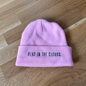 Rosa mössa - Ariana Grande merch - Säljer en ljusrosa stickad mössa med svart broderad text 'HEAD IN THE CLOUDS' på framsidan och 'ARIANA GRANDE' på baksidan. Äkta Ariana Grande merch från the honeymoon tour. Jättebra kvalité!