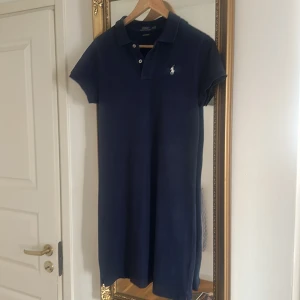 Marinblå pikéklänning från Polo Ralph Lauren - Klassisk marinblå pikéklänning från Polo Ralph Lauren med korta ärmar och krage. Klänningen har knappar framtill och den ikoniska loggan broderad på bröstet. Perfekt för en sportig och stilren look.