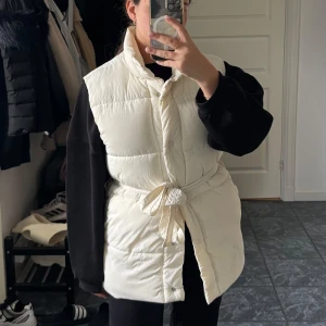 Vit dunväst med knytband - Snygg och trendig vit dunväst med knappar framtill och knytband i midjan. Västen har en hög krage och en något längre passform som ger en cool siluett. Perfekt att slänga över en hoodie för en avslappnad look.