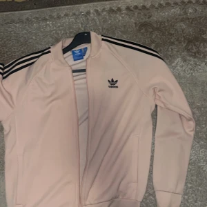 Ljusrosa Adidas originals tröja - Säljer en ljusrosa Adidas track jacket med svarta ränder på ärmarna och Adidas-logga på bröstet. Jackan har dragkedja framtill och är långärmad. Klassisk sportig stil och bekväm passform.