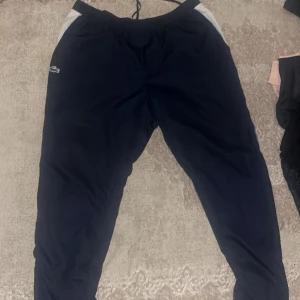 Marinblå träningsbyxor från Lacoste Sport - Säljer ett par marinblå träningsbyxor från Lacoste Sport med vita och svarta detaljer längs sidorna samt orange Lacoste-tryck. Byxorna har resår i midjan och snörning för bästa passform. Perfekta för sportiga aktiviteter.