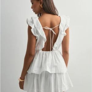 Vit volangklänning med öppen rygg och knyt - Supersöt vit kortklänning med volangdetaljer och omlott framtill. Klänningen har öppen rygg med knytband och är ärmlös med volangkant. Perfekt för sommarens alla tillfällen! Endast testad helt ny 🥰 