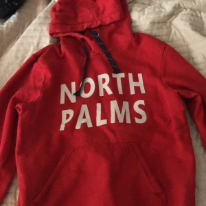 Röd hoodie med tryck från North Palms - Röd hoodie med vit text 'NORTH PALMS' på bröstet och blå/vita snören i huvan. Stor känguruficka framtill och mjukt material. Perfekt för en avslappnad stil.