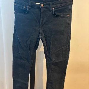 Svarta jeans från Nudie Jeans - Säljer ett par svarta jeans från Nudie Jeans med klassisk femficksdesign och knappgylf. Jeansen har en rak passform och är tillverkade i ekologisk bomull. Perfekta för dig som gillar stilrena och tidlösa byxor.