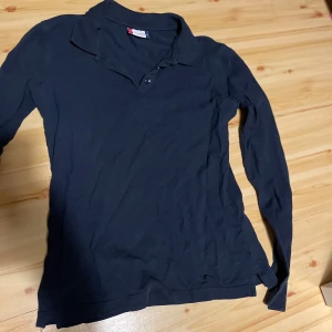 Mörkblå pikétröja från Clique - Säljer en mörkblå långärmad pikétröja från Clique. Tröjan har klassisk krage och knäppning framtill. Perfekt för en stilren och enkel look. Det står strlk m men den passar mer xs