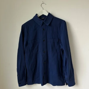 Mörkblå Arket overshirt med knappar - Säljer en stilren mörkblå overshirt med klassisk krage och stora bröstfickor. Skjortan har långa ärmar och stängs med svarta knappar framtill. Perfekt att bära som lager över en t-shirt eller tröja. Artnr: Ö269