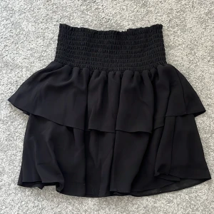 Svart volangkjol från Chelsea - Supersöt svart kjol från Chelsea med bred smock i midjan och dubbla volanger. Kjolen är kort och har en flowy känsla som gör den extra bekväm och snygg. Perfekt att styla med en enkel topp eller stickad tröja.