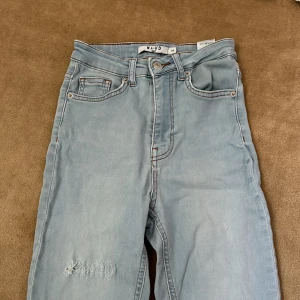 Ljusblå slitna jeans från NA-KD - Säljer ett par ljusblå jeans från NA-KD med slitna detaljer på benen. Jeansen har klassisk femficksdesign, hög midja och raka ben. Perfekta för en avslappnad och trendig look.