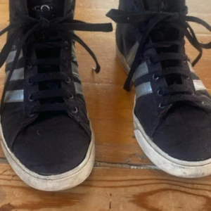 Svarta höga sneakers med silvriga ränder - Säljer ett par svarta höga sneakers från Adidas med klassiska silvriga ränder på sidorna. Skorna har snörning och vit sula, perfekt för en sportig och avslappnad stil. Passar dig som gillar streetwear och vill ha något bekvämt till vardags.