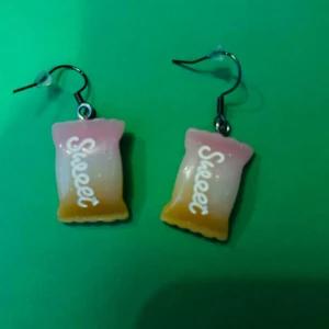 Söta örhängen med godispåse-design - Ett par unika örhängen formade som små godispåsar i rosa, vitt och gult med texten 'Sweet' på framsidan. Perfekta för dig som gillar lekfulla accessoarer och vill sticka ut lite extra.