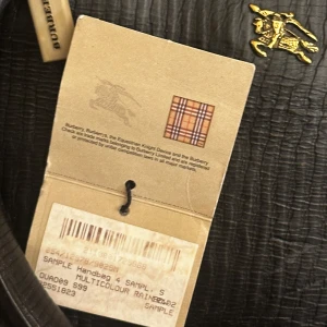 Svart handväska från Burberry - Säljer en svart handväska från Burberry med guldfärgade metalldetaljer och strukturmönstrad yta. Väskan har dubbla handtag och en dekorativ rem i guld. Insidan har fack med dragkedja och Burberrys klassiska rutiga etikett.