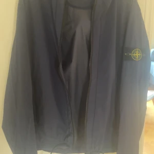 Blå vindjacka från Stone Island - Säljer en blå vindjacka från Stone Island med huva och dragkedja. Jackan har den klassiska loggan på ärmen och är tillverkad i ett lätt syntetmaterial. Perfekt för blåsiga dagar och enkel att matcha med olika outfits.
