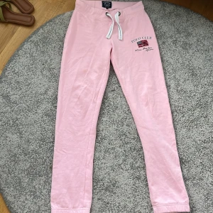 Ljusrosa mjukisbyxor från VNSOA Polo Club - Säljer ett par ljusrosa mjukisbyxor från VNSOA Polo Club med dragsko i midjan och broderad logga samt amerikansk flagga på ena benet. Byxorna har ribbade muddar vid bensluten och är perfekta för en avslappnad stil.