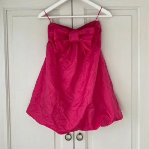 Rosa axelbandslös klänning med rosett - Supersöt rosa kortklänning som är axelbandslös och med stor rosett framtill. Ballongformad nederdel och smockad rygg. Endast annvänd en gång så jättebra skick🌟 Hon på sista bilden är 170cm, passar även kortare och lite längre. Perfekt nu i sommar! 